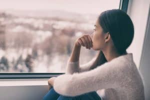 Une femme regarde par la fenêtre un paysage enneigé, dépression hivernale saisonnière